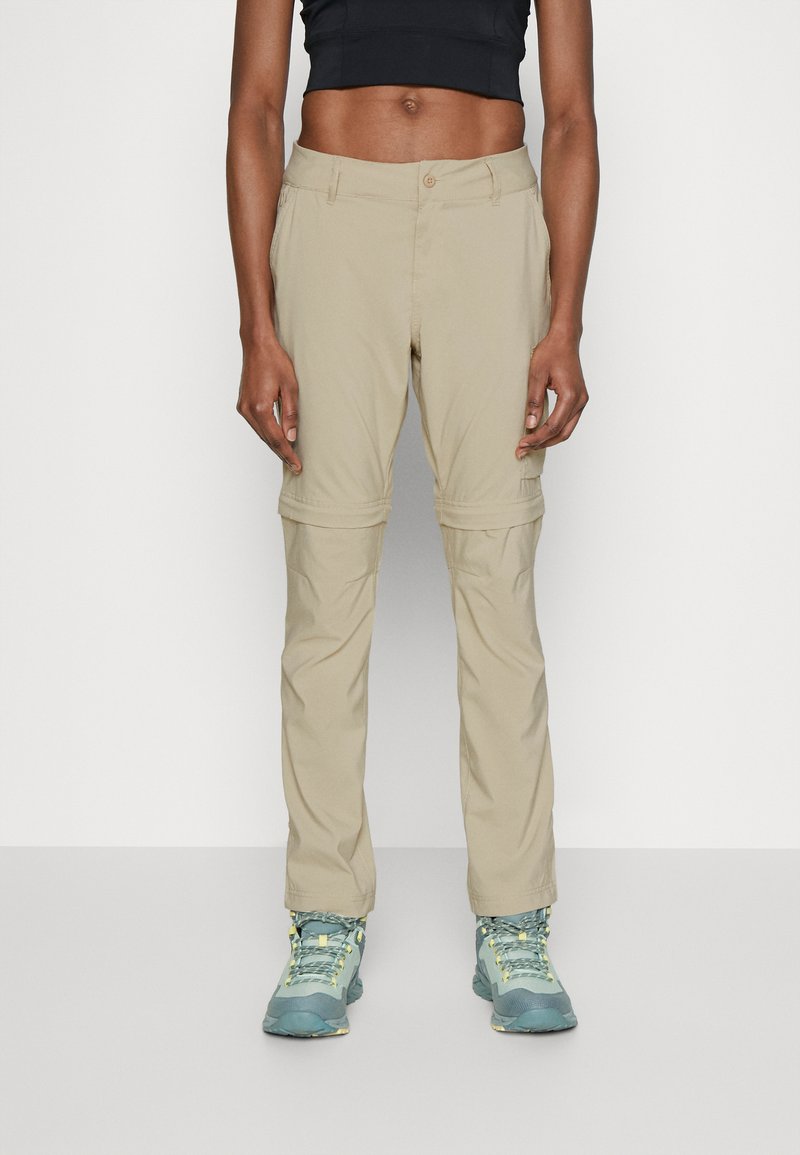 Beige Zip-Off-Hosen mit schmalem Schnitt und Knopfverschluss, aus leichtem Stoff, kombiniert mit blauen und gelben Wanderschuhen.