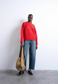 Femme portant un pull rouge, un jean à jambes larges bleu, des chaussures noires à motifs, tenant un grand sac beige, debout contre un mur blanc.