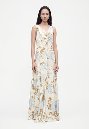 Femme aux longs cheveux noirs portant une robe maxi blanche sans manches avec des motifs floraux bleus et beiges, debout devant un fond blanc uni.