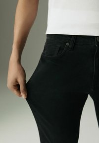 Zwarte denim jeans met een slim fit, een enkele voorkantzak en contrasterende stiksels. Een hand grijpt de stof bij de dij.