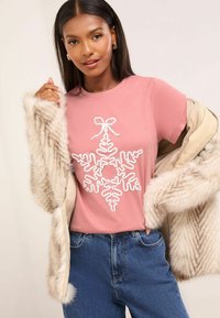 Camiseta rosa de manga corta con un diseño de copo de nieve blanco, llevada debajo de un abrigo de piel sintética beige claro con patrones de rayas.