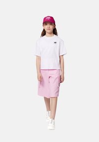 T-shirt blanche raccourcie avec logo noir, shorts cargo rose clair avec poches latérales et baskets blanches. Casquette rose avec design de logo détaillé.