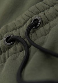 Gros plan sur un tissu vert olive avec un cordon noir passé à travers des œillets métalliques sur une taille froncée.
