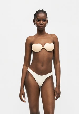 Mujer con piel oscura y cabello trenzado lleva un sujetador beige sin tirantes y braguitas de bikini a juego de corte alto, de pie frente a un fondo claro y liso.