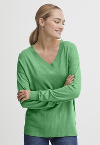 Pulover verde din tricot cu guler în V, mâneci lungi și croială lejeră. Textură moale și detalii subtile la tiv. Asortat cu blugi albaștri.