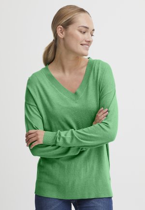 Groene gebreide trui met een V-hals, lange mouwen en een ontspannen pasvorm. Zachte textuur en subtiele zoomdetails. Gecombineerd met blauwe jeans.