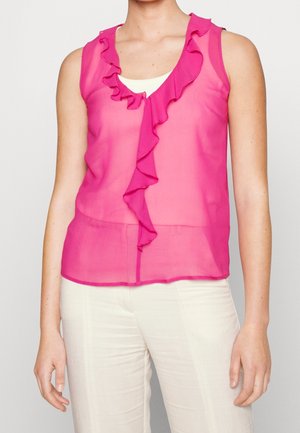 Hihaton läpikuultava vaaleanpunainen pusero, jossa röyhelöt edessä, päällä valkoinen toppi ja valkoiset housut.