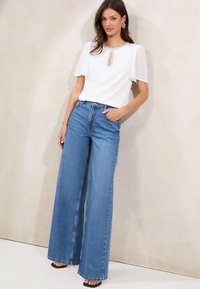 Weißes Kurzarmoberteil mit Schlüssel-Ausschnitt und verziertem Dekolleté, kombiniert mit hochtaillierten, weit geschnittenen blauen Jeans. Schwarze Sandalen.