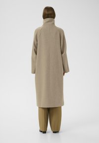 Cappotto in lana beige con colletto alto, maniche lunghe e taglio dritto, caratterizzato da un design della schiena senza cuciture e una texture minimalista.
