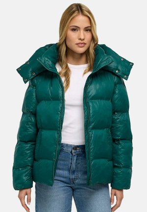 Femme portant une doudoune vert foncé avec un grand col, une chemise blanche et un jean bleu sur un fond clair uni.