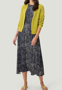 Cardigan en tricot jaune sur une robe bleu marine avec un motif tourbillonnant blanc et doré. Le modèle porte des sandales à glissière dorées. Texture de tissu lisse.