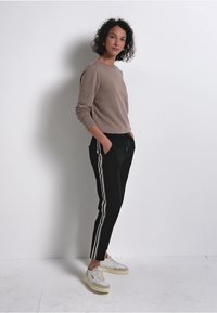 Beige sweatshirt, svarta byxor med vita sidostråk, vita och beige sneakers. Modellen står mot en enkel vit bakgrund.