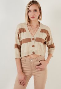 Krémový zkrácený cardigan s hnědými pruhy, ribbovanou texturou, výstřihem do V a čtyřmi velkými hnědými knoflíky. Stylizováno s krémovými úzkými kalhotami.