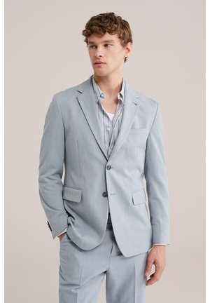 Uomo con abito sartoriale grigio chiaro composto da giacca con due bottoni e pantaloni abbinati, abbinato a una camicia a righe e una maglietta bianca sotto.