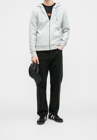 Grauer Zip-Hoodie mit Kapuze, darunter ein schwarzes T-Shirt, schwarze Jeans mit geradem Bein, schwarze Schuhe mit weißen Streifen und eine schwarze Kappe.