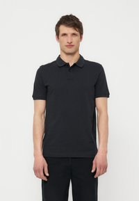 Uomo in piedi, rivolto verso di noi, con polo nera a maniche corte e pantaloni neri su uno sfondo bianco uniforme.