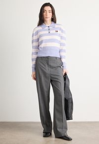 Pulover tricotat cu dungi mov și roz, cu guler și nasturi, combinat cu pantaloni gri cu dungi. Geantă din piele în mână.