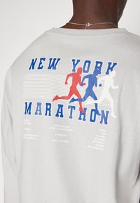 Lys grå langermet skjorte med "NEW YORK MARATHON" i blå tekst. Grafikk av løpefigurer i rødt, blått og hvitt. Tekst detaljer under.