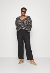 Top negro con estampado, escote en V y corte holgado, combinado con pantalones anchos plegados negros y sandalias de tacón beige. Las gafas de sol realzan el conjunto.