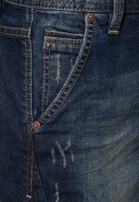 Jeans de mezclilla azul oscuro con una textura desgastada, costuras en dorado contrastante y detalles de hardware en cobre en la zona de los bolsillos.