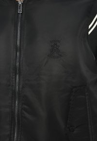 Fekete bomberdzseki cipzáras záródással, hímzett logóval a mellkason, oldalzsebekkel és kontrasztos, csíkos válldíszítéssel.