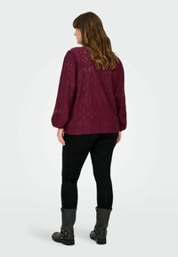 Bordo čipkasta bluzica z dolgimi napihnjenimi rokavi z cvetličnim vzorcem, nosena nad črnimi oprijetimi kavbojkami in s sivimi čevlji do kolen.