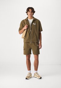 Olijfgroen zip-up jack en bijpassende shorts van textuurstof. Witte t-shirt eronder, met een tan crossbody tas. Beige sneakers.