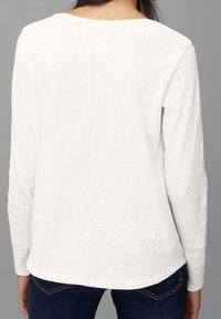 Vrouw die een eenvoudig wit shirt met lange mouwen en donkerblauwe spijkerbroek draagt, gezien van achteren tegen een grijze achtergrond.