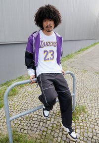 Lilla og sort vindjakke over en hvid Lakers t-shirt, grå træningsbukser med hvide detaljer, og sort-hvide sneakers, siddende på et gelænder.