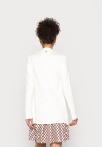 Blazer blanco de mangas largas y corte estructurado, con un pequeño detalle dorado en el cuello, combinado con una falda marrón estampada.