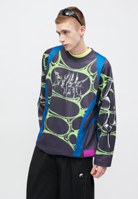 Φούτερ με μακριά μανίκια σε σκούρο μωβ χρώμα με πράσινο αφηρημένο μοτίβο και μπλε λεπτομέρειες· διαθέτει λογότυπο και υφή υφάσματος. Φοράει μαύρο παντελόνι.