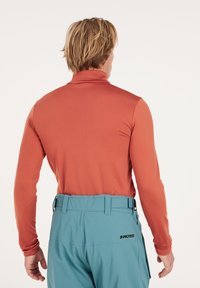 Lange mouwen, hoge hals top in verbrande sinaasappel, gecombineerd met lichtblauwe broek. Stof lijkt glad met een getailleerd ontwerp. Logo op de tailleband.