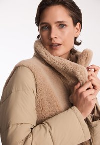 Beżowa kurtka puchowa z puszystym, karmelowym kołnierzem z syntetycznego futra. Materiał jest gładki o miękkiej fakturze, ukazując nowoczesny, przytulny projekt warstwowy.