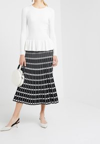 Valkoinen ribattu pitkähihainen paita, jossa on peplum-helma, yhdistettynä musta-valkoiseen laskostettuun hameeseen, jossa on ruutukuviota. Mukana pyöreä valkoinen laukku.
