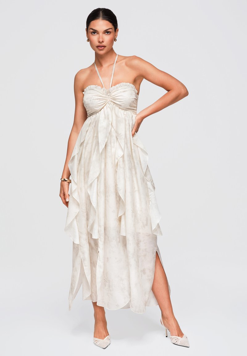 Femme portant une robe bustier beige clair avec un corsage froncé et des couches à volants, associée à des chaussures blanches à talons aiguilles pointues et des boucles d'oreilles créoles dorées.