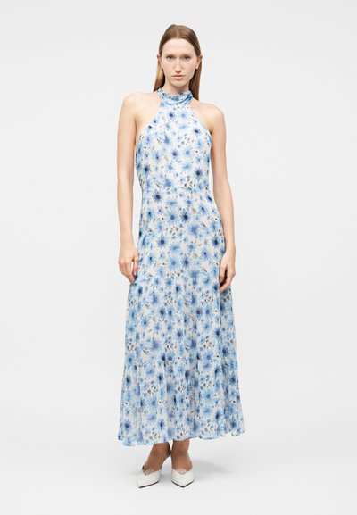 Γυναίκα που στέκεται, φορώντας ένα μακρύ φόρεμα με λουλουδάτο σχέδιο σε γαλάζιο χρώμα και λευκά μυτερά τακούνια, μπροστά σε ένα καθαρό λευκό φόντο.