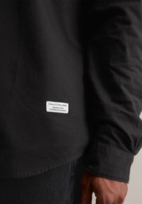 Črna dolga majica z dolgimi rokavi iz bombaža s poševnim robom. Ima belo oznako z blagovno znamko in besedilom na spodnjem levem delu. Gladka tekstura.