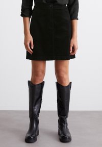 Jupe mini en velours côtelé noir avec une silhouette ajustée, fermeture à boutons sur le devant et deux poches latérales. Associée à des bottes en cuir noir jusqu'aux genoux.