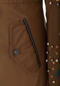 Braune Jacke mit einer Seitentasche, die eine schwarze Borte und einen Knopf hat. Der Stoff enthält Metallnieten und gestickte Akzente in verschiedenen Formen.