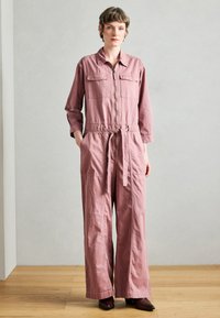 Világos rózsaszín hosszú ujjú overál elülső zsebekkel, cipzáras lezárással és övvel a deréknál. Széles, légies nadrágszárak és texturált anyag.