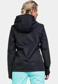 Frau mit blonden Haaren im Dutt, die eine schwarze Kapuzenjacke und hellblaue Hosen trägt, von hinten vor einem schlichten Hintergrund gezeigt.