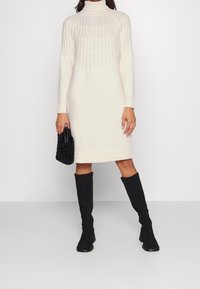 Abito lavorato a maglia color crema con collare alto e parte superiore testurizzata, abbinato a stivali neri alti fino al ginocchio e una piccola borsa nera testurizzata.