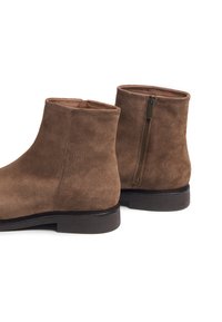 Bottines en daim marron avec une texture lisse, bout rond et fermeture éclair sur le côté. La semelle est sombre et légèrement surélevée.