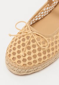 Bežové tkané espadrilky s mriežkovaným vzorom, guľatým tvarom, ozdobnou mašľou na prednej strane a prírodnou jutovou podrážkou. Plochý dizajn s hladkou textúrou.