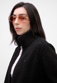 Svart fleecejakke med høy krage. Modellen har på seg rosa fargede solbriller. Festet skjorte synlig under, med en liten trykt detalj.