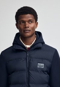 Puffjacka i marinblått med huva, med en quiltad design, dragkedjor och en "POLO CLUB" logotyp på bröstet. Strukturerad tyg på ärmarna.