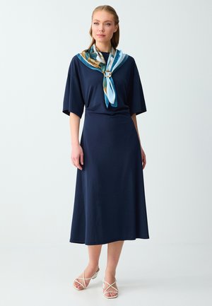 Femme portant une robe midi bleu marine, une écharpe multicolore avec un anneau, et des sandales blanches à talons avec des brides, debout sur un fond blanc.