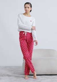 Λευκό μακρυμάνικο τοπ με μικρή λεπτομέρεια κόκκινης καρδιάς, σε συνδυασμό με κόκκινο παντελόνι με λευκά σχέδια καρδιών. Μαλακό ύφασμα, χαλαρή εφαρμογή.
