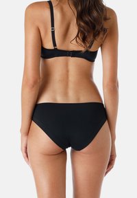 Donna vista da dietro che indossa un reggiseno nero con spalline regolabili e coordinato con un perizoma nero in stile bikini, su uno sfondo neutro.