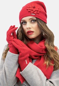 Czerwona dziana czapka beanie z detalem w postaci filcowego kwiatu, pasujące czerwone rękawiczki i szalik. Miękka tekstura i elegancki design; wszystko w jaskrawoczerwonym kolorze.